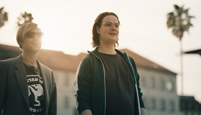 2 junge lachende Männer stehen nebeneinander, einer mit blonden Haaren und Seitenscheitel, der andere mit langen dunklen Haaren.