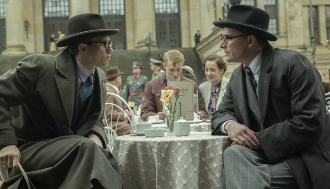 Zwei junge Männer mit Anzug und Krempenhut sitzen sich dicht nach vorne gebeugt an einem Café-Tisch gegenüber