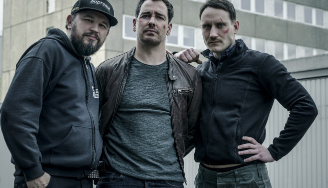 Foto zu "Dogs of Berlin". von links nach rechts drei Männer, einer mit Schirmmütze, einer mit Bart und einer ohne Bart.