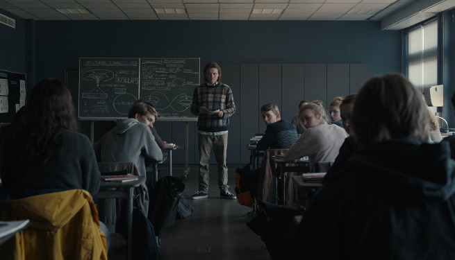 Foto aus "Dark". In einem dunklen Klassenraum steht vorne eine langhaarige Person mit Zettel in der Hand, einige Schüler in den Reihen drehen sich nach hinten um.