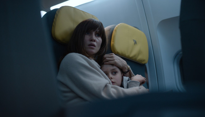 Eine junge Frau mit dunklen Haaren und Pony sitzt in einem Flugzeug und hält ihren Sohn eng im Arm, ihr Mund ist leicht geöffnet und ihr Blick angsterfüllt