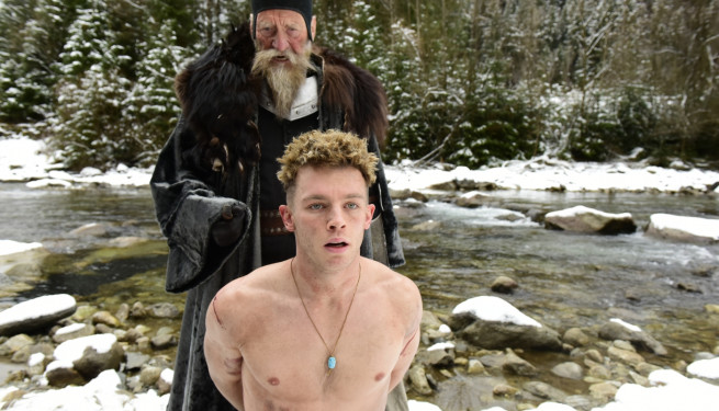 Foto aus "Narziss und Goldmund". Ein junger Mann kniet mit nacktem Oberkörper und nach hinten verschränkten Armen auf dem Boden, hinter ihm ein Geistlicher mit schwarzem Umhang und langem Bart.