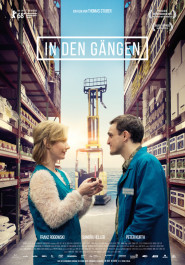 Filmplakat zu "In den Gängen". In einer Lagerhalle zwischen meterhohen Regalen stehen sich ein Man und eine Frau in blauer Kleidung gegenüber.