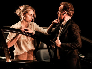 Eine blonde Frau schaut einen kurzhaarigen Mann auffordernd an und greift ihm ans Kinn.